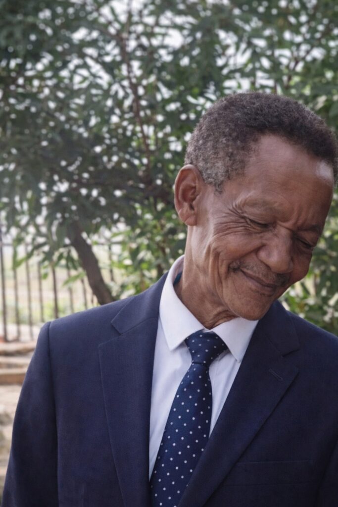 Tah Magic wearing a navy formal suit and tie, smiling gently outdoors, representing wisdom and guidance from a respected elder in a South African family